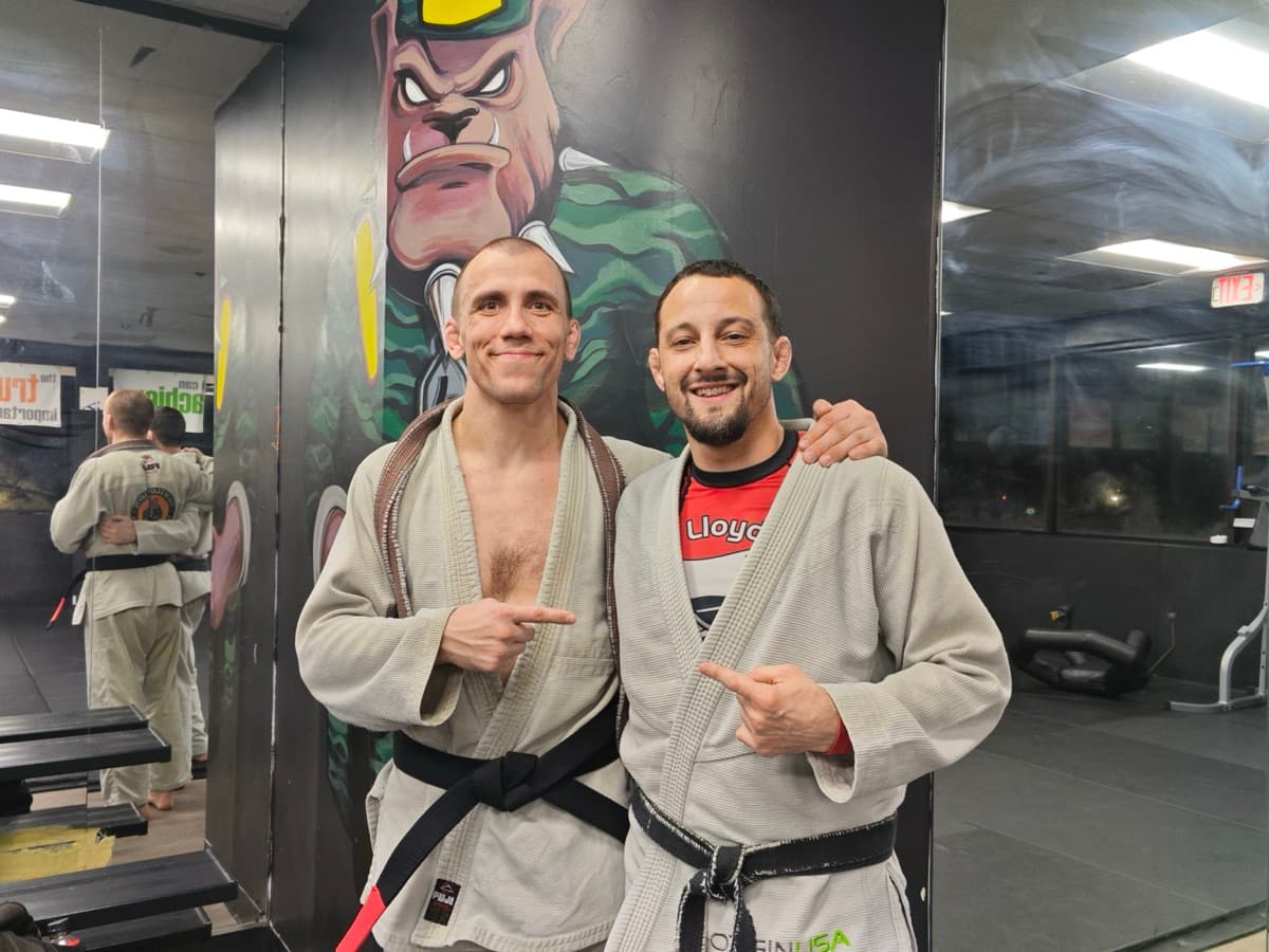 Leo and a training partner both in white gi with black belts, pointing at each other in front of a BJJ gym mural