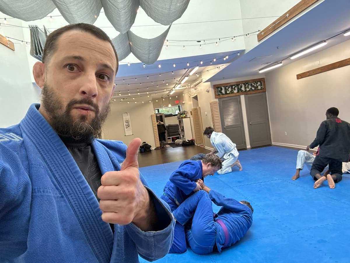 Leo in a blue gi giving a thumbs up selfie with students actively training on the mats behind him