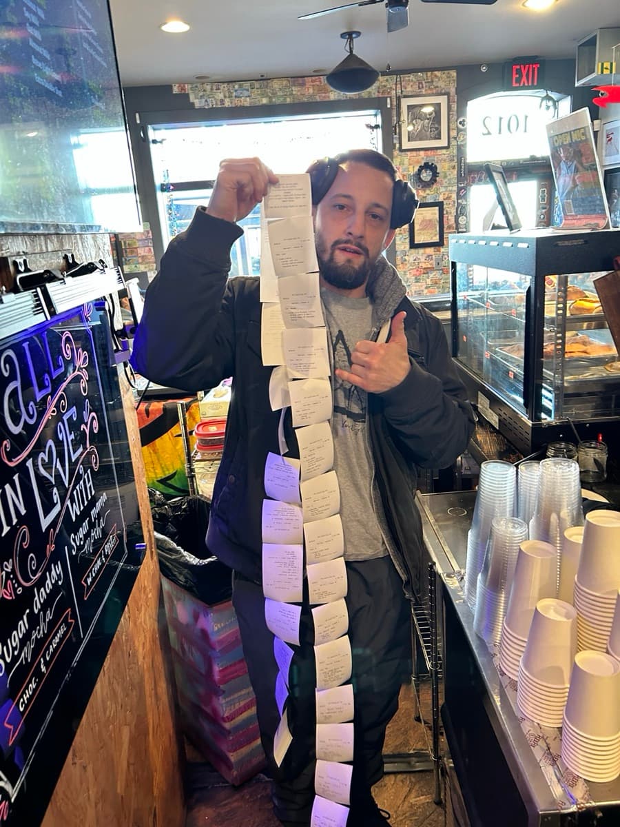 Leo holding a long receipt tape of customer orders while smiling with a shaka sign inside a coffee shop