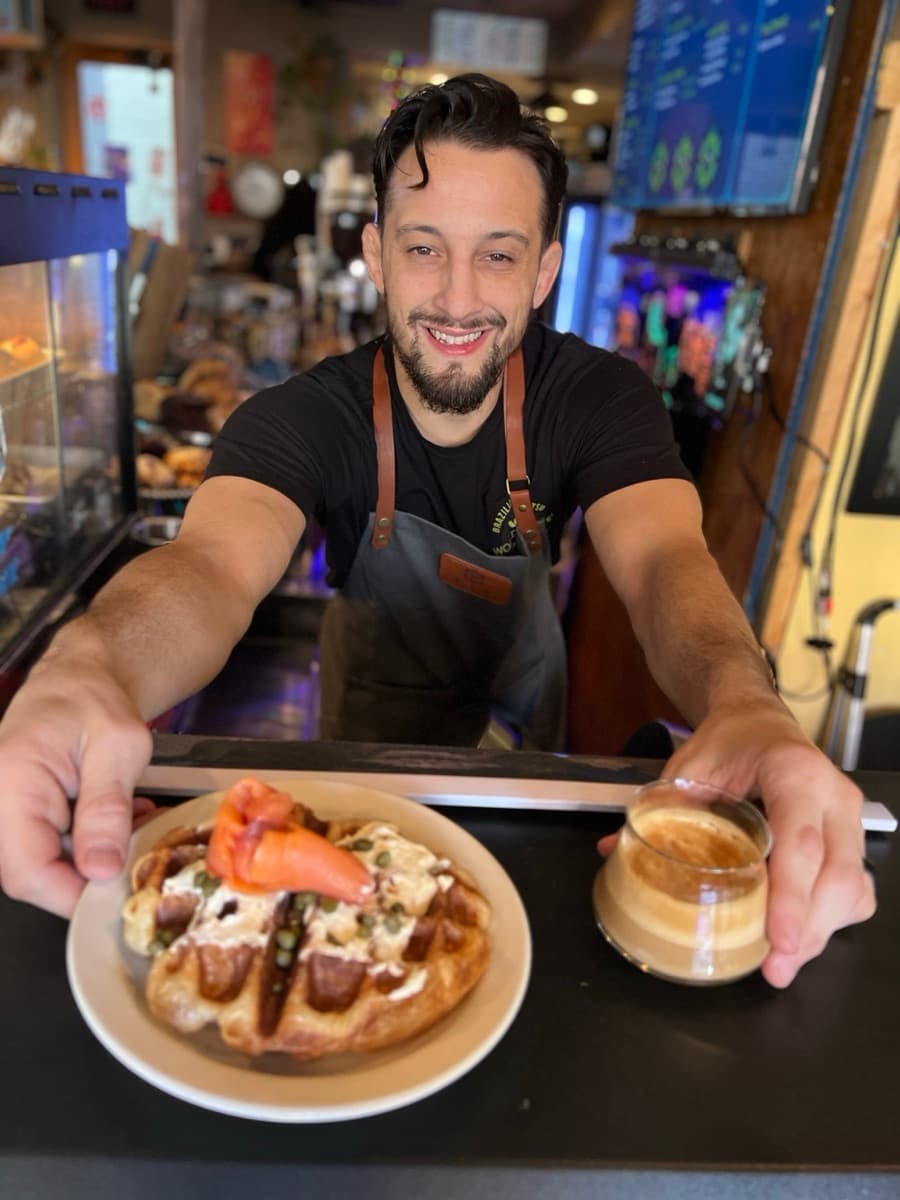 Leo in an apron leaning over a counter presenting a beautifully plated dish and espresso with a warm smile